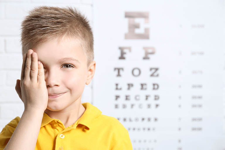 A young boy with blond hair in a yellow shirt covers one eye with his hand, embodying the essence of children's eye tests while standing in front of a blurred chart. His slight smile suggests a playful or comfortable visit to the optometrist.