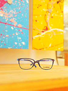 A pair of eyeglasses rests on a wooden surface, with vibrant abstract art in blue and yellow hues hanging on the wall behind. This calming scene is perfect for children’s eye tests, ensuring a comforting ambiance for young ones.