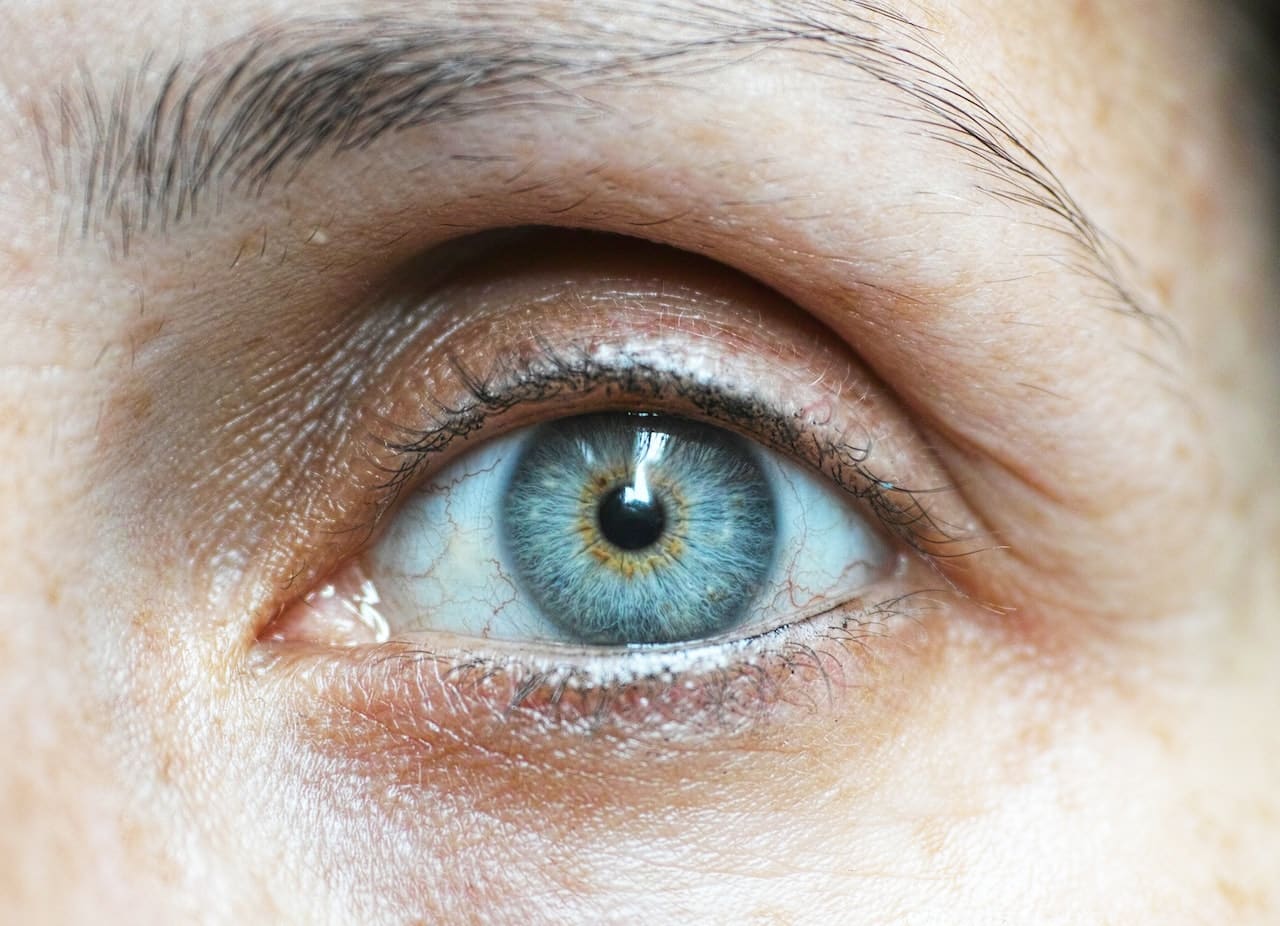 Close-up of a human eye with a blue iris and visible details such as eyelashes and surrounding skin texture. The eye is open, showing a clear view of the intricate pattern and color variations within the iris.