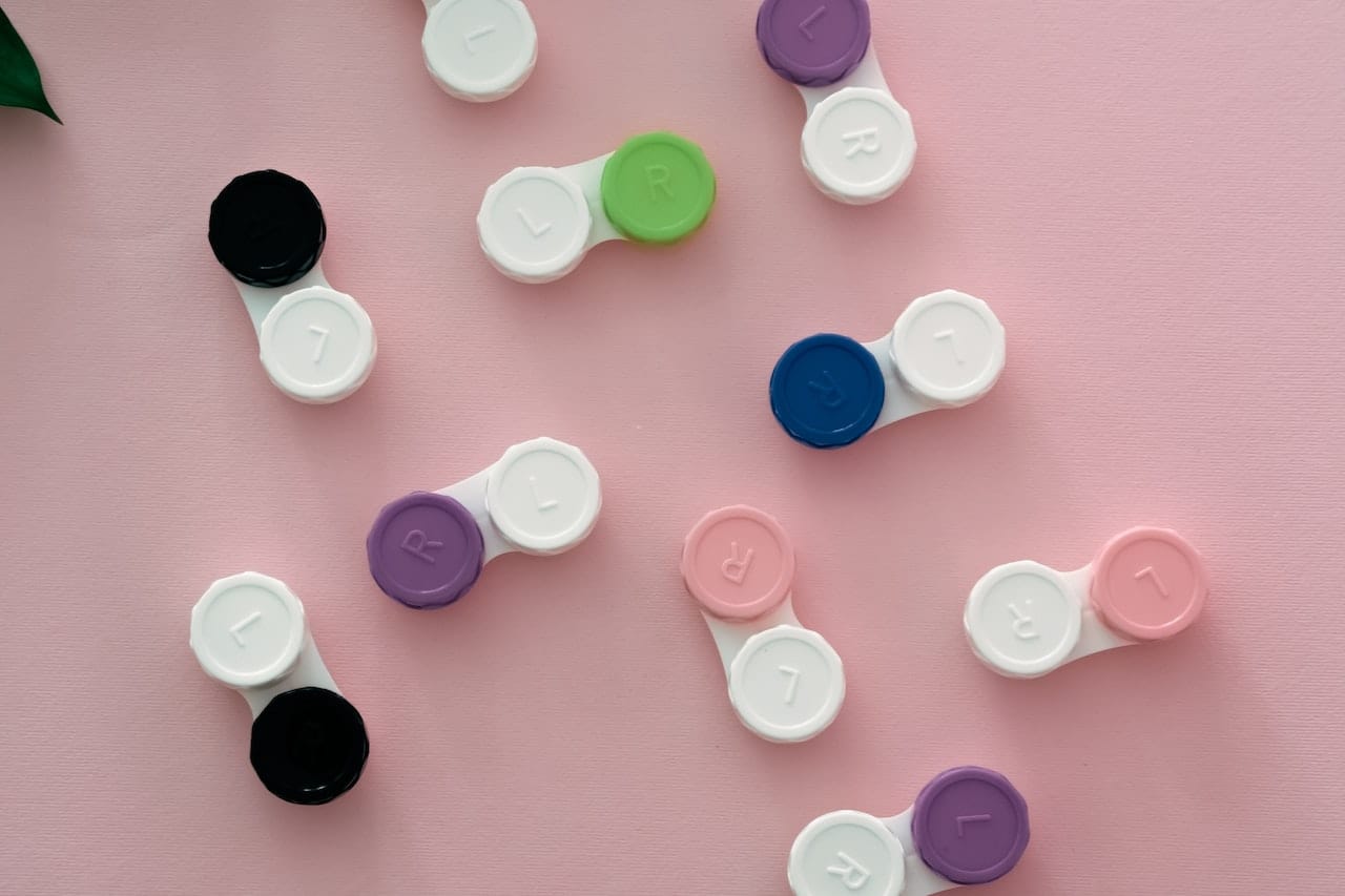 Contact lens cases in various colors, including black, purple, green, blue, and pink, are scattered on a pink surface. Each case is labeled with "L" and "R" for left and right.