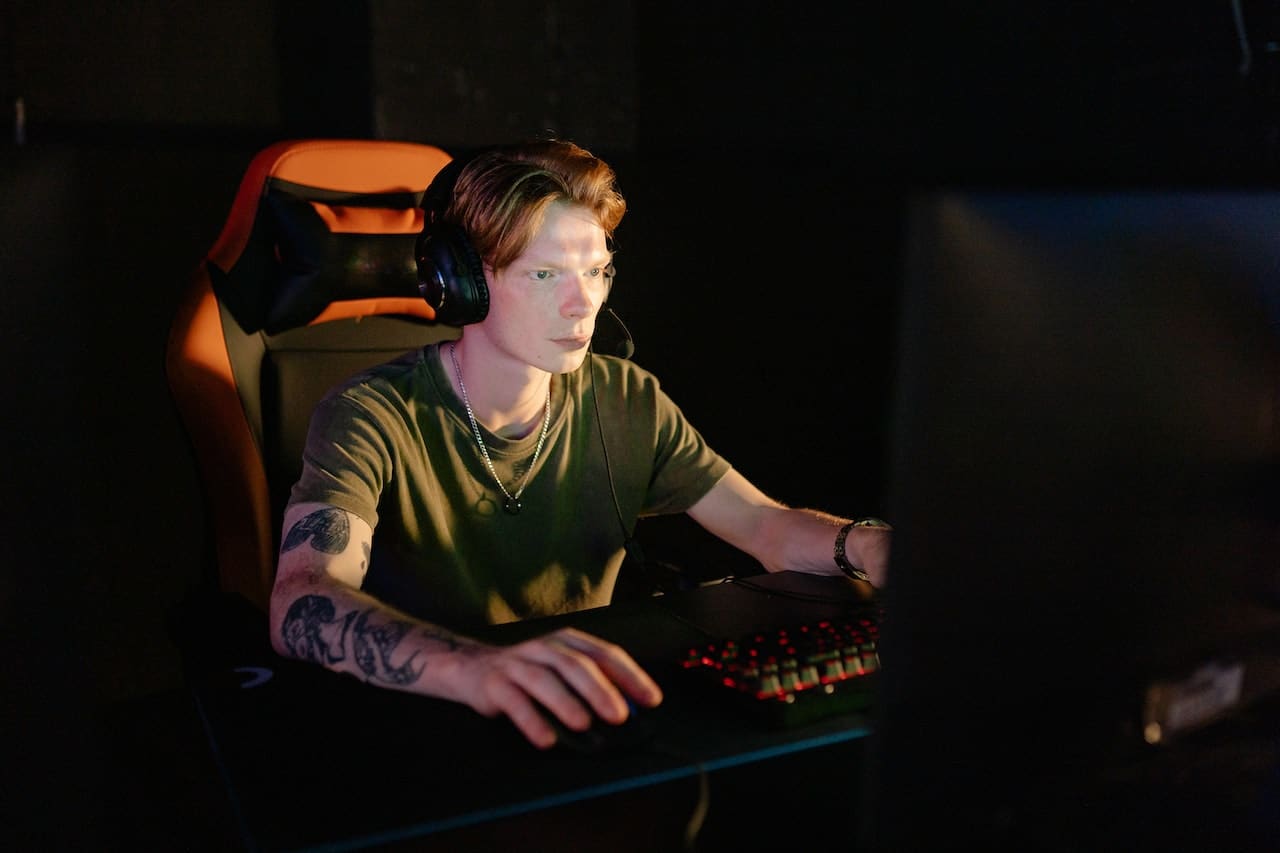 A person with a tattoo on their arm is sitting in an orange gaming chair, wearing headphones, and focused on a computer screen in a dimly lit room. They are using a keyboard and mouse.