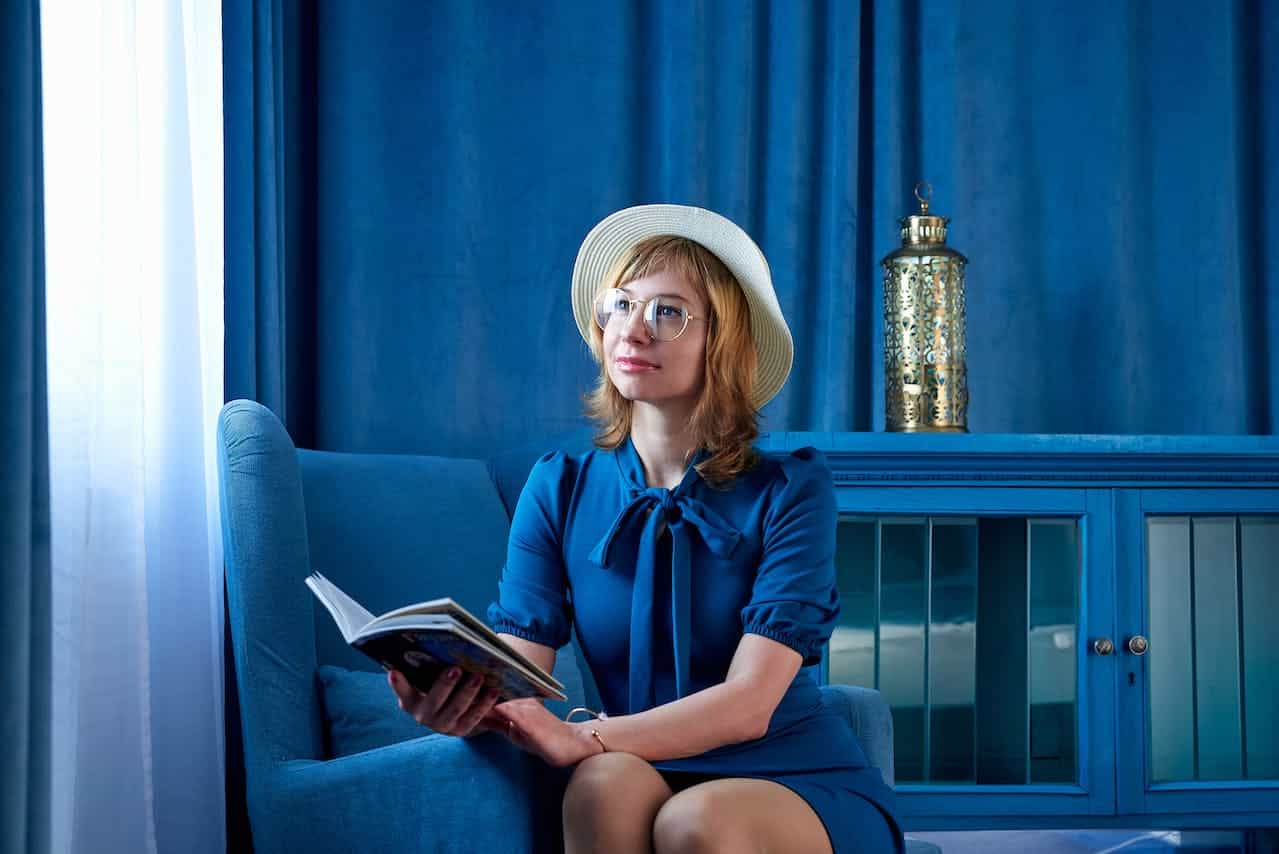 A woman wearing a blue dress and straw hat sits in a blue armchair, holding an open book. She is looking thoughtfully out a window, with a blue cabinet and ornate golden lantern in the background.