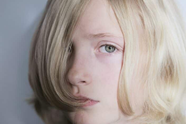 A close-up of a young person with long, light blonde hair covering part of their face. They have fair skin and are gazing directly at the camera against a plain background.