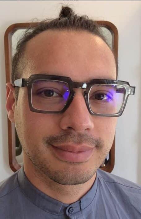 A person with glasses reflecting their visit to opticians Camden has slight facial hair and hair tied up as they smile slightly in front of a mirror They're donning a blue gray collared shirt against a light colored wall backdrop