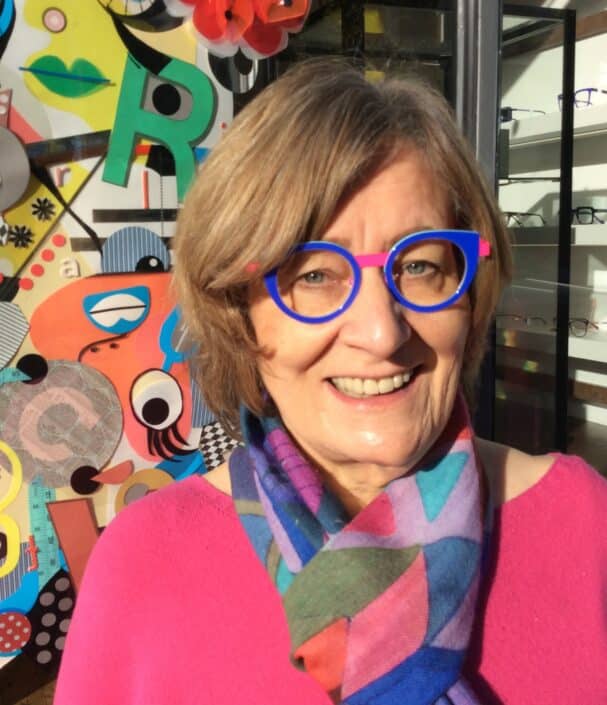 A woman with short hair is wearing bright blue glasses and a multicolored scarf smiling outside an optician's shop in Camden The vibrant backdrop features various shapes and patterns echoing the artistic flair of this lively neighborhood