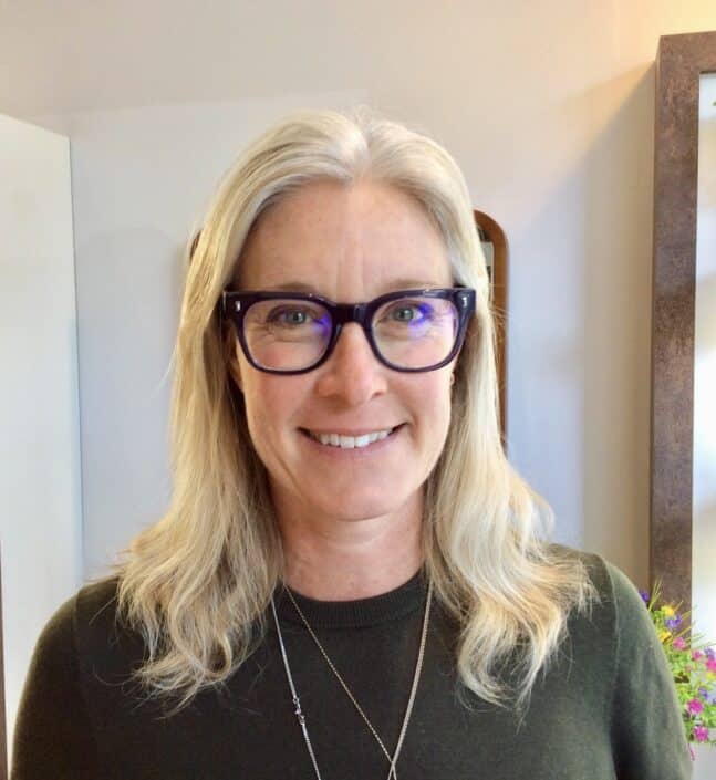 A woman with long blonde hair is smiling while wearing glasses possibly from an optician in Camden She is dressed in a dark green top and stands indoors with a mirror and white wall accentuating her joyful expression