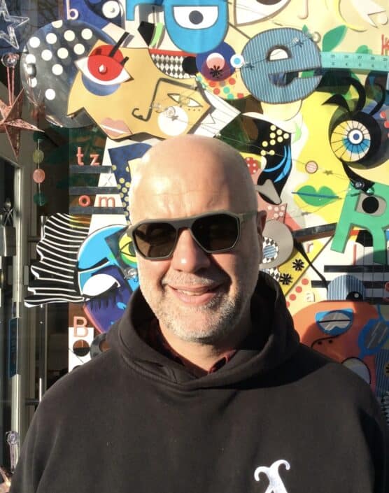 In the radiant sunshine a person wearing sunglasses and a dark hoodie stands smiling in front of a colorful abstract mural brimming with shapes letters and faces—perhaps they just came from an optician's visit in Camden
