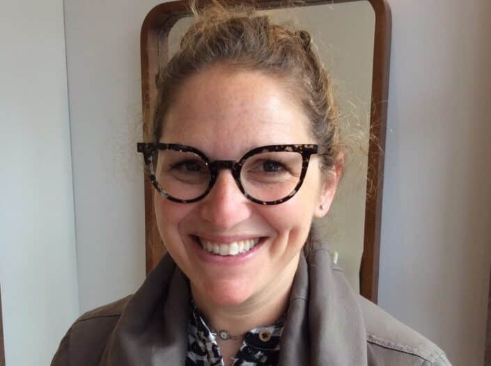 A person with curly hair smiles warmly sporting round eyeglasses from a stylish opticians in Camden Dressed in a jacket and patterned shirt they stand indoors with a mirror reflecting their fashionable look in the background