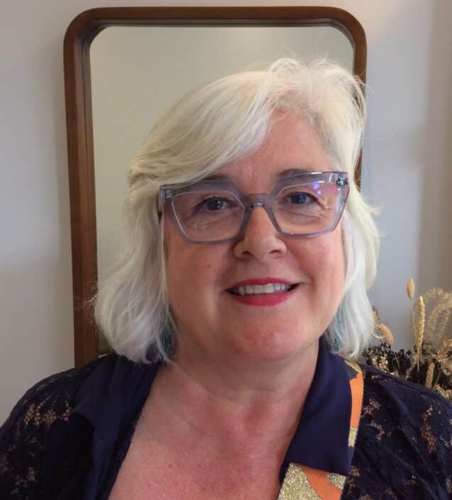 A woman with short white hair and clear framed glasses smiles at the camera She's dressed in a navy top standing in front of a brown framed mirror adorned with decorative plants—a nod to chic style you might find at opticians in Camden