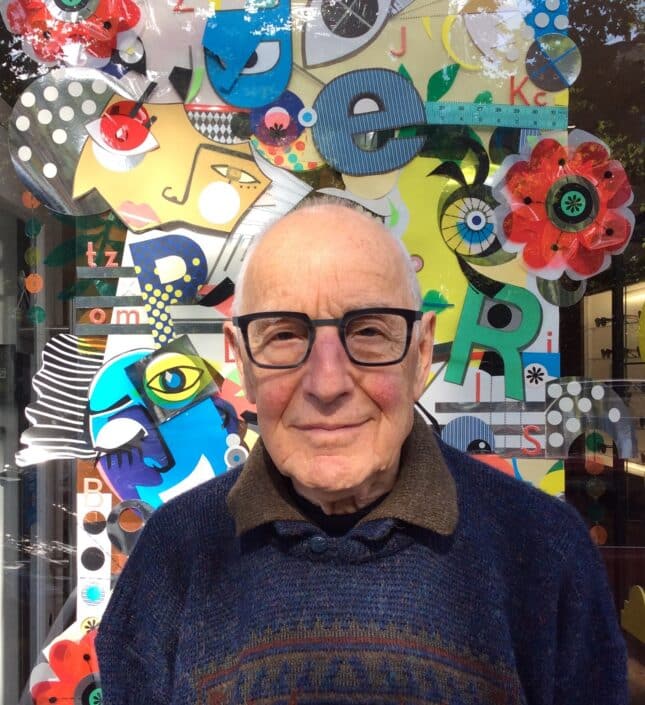 An elderly person with glasses and a dark sweater stands in front of a colorful abstract mural in Camden adorned with various shapes patterns and letters