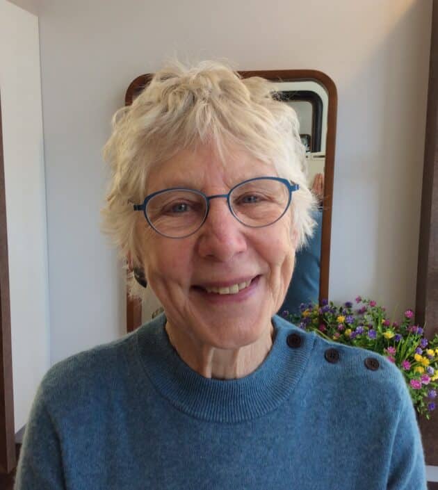 An elderly person with short gray hair and blue glasses possibly from a local opticians in Camden smiles warmly at the camera They wear a blue sweater with button details In the background there are flowers and a mirror reflecting their cheerful demeanor