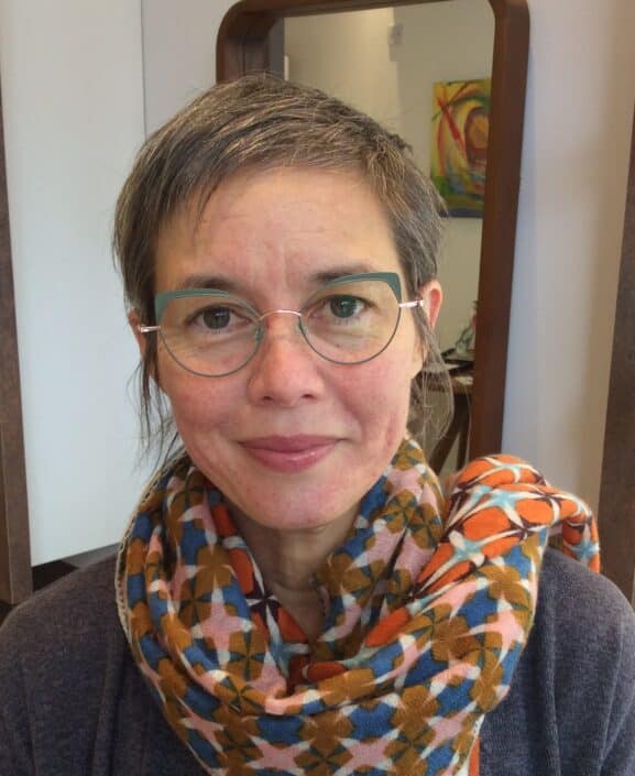 A person with short gray hair and glasses perhaps from opticians in Camden smiles at the camera They sport a colorful scarf with geometric patterns over a dark top In the background a mirror reflects an abstract painting