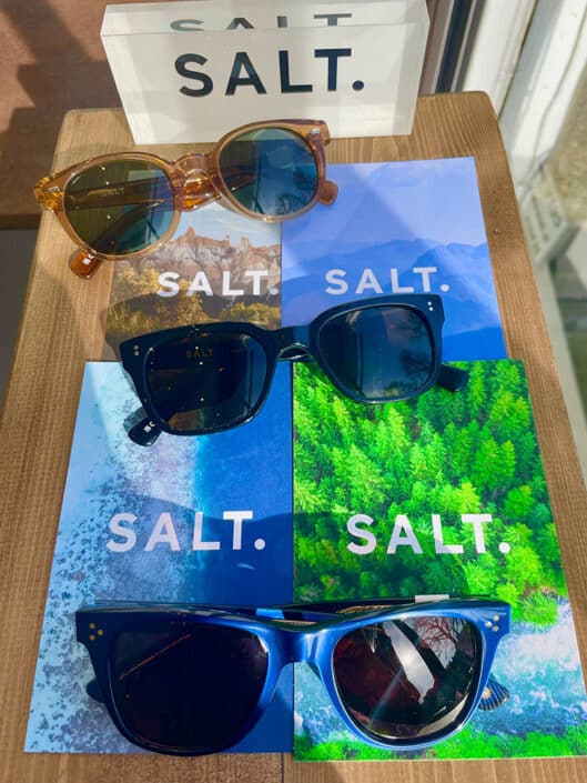 Three pairs of SALT sunglasses are displayed on a wooden surface each resting on vibrant nature themed backdrops with images of mountains ocean and forests A stylish SALT sign enhances the lively scene in the background