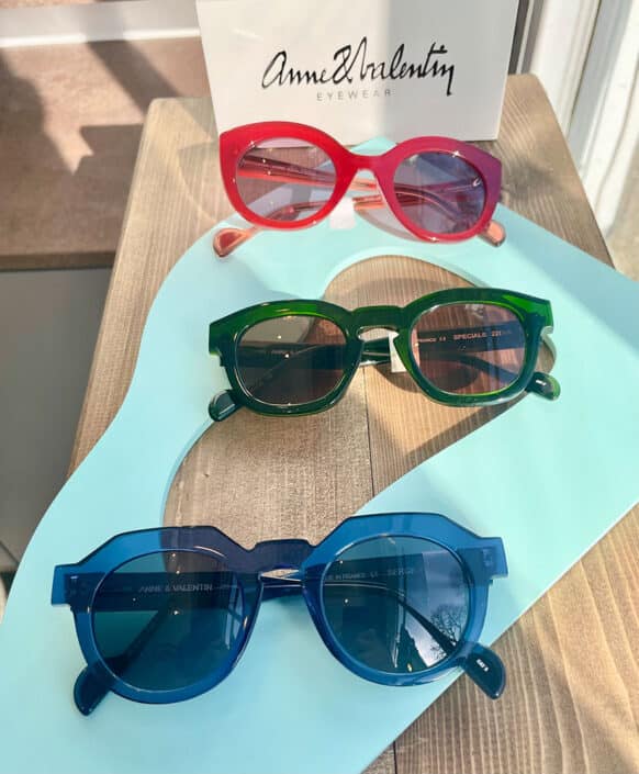 Three vibrant pairs of Anne et Valentin sunglasses—red cat eye green round and blue square frames—adorn a light blue display on a wooden surface near a window A branded paper bag sits gracefully in the background enhancing the chic allure of these sunglasses